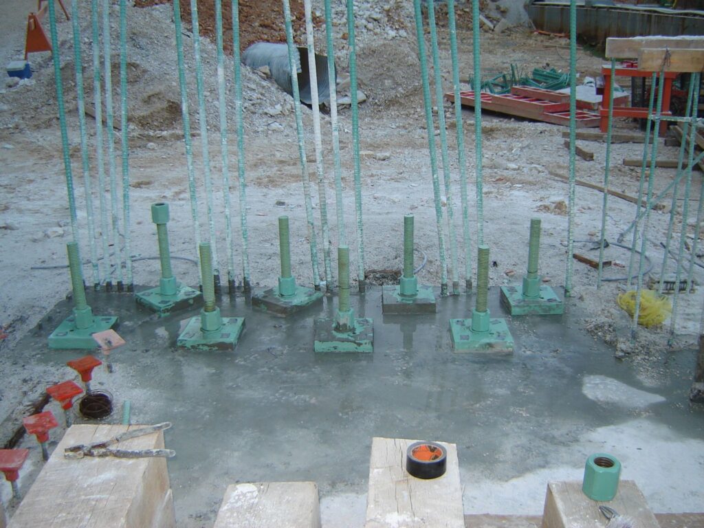 Construction site showing green foundational steel columns and rebar rods embedded in concrete. Various tools and building materials are scattered around on the ground.