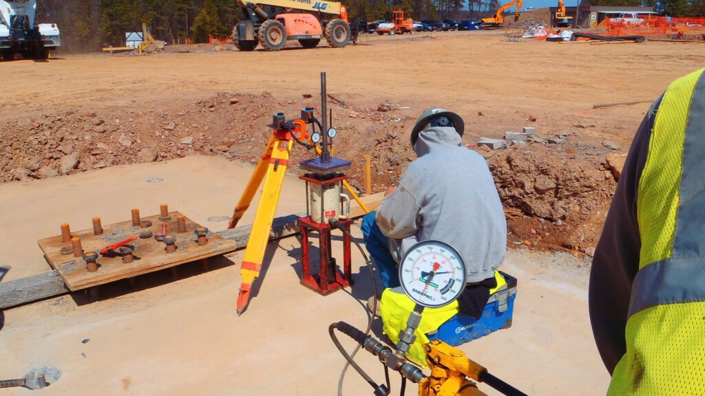 A construction worker in a gray hoodie operates machinery on a construction site. The equipment measures pressure, with a gauge and other instruments visible. The sandy site has construction vehicles and materials in the background.