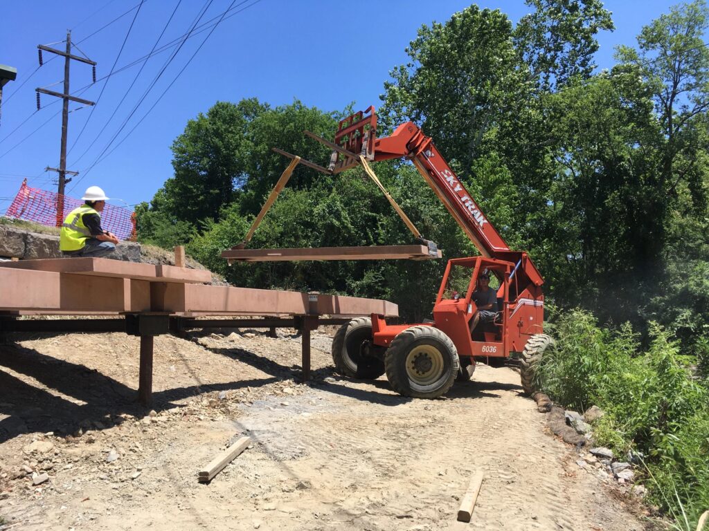 A construction worker in a white hard hat observes as a red forklift moves a steel beam on a dirt path. The area is surrounded by green trees and electric poles.