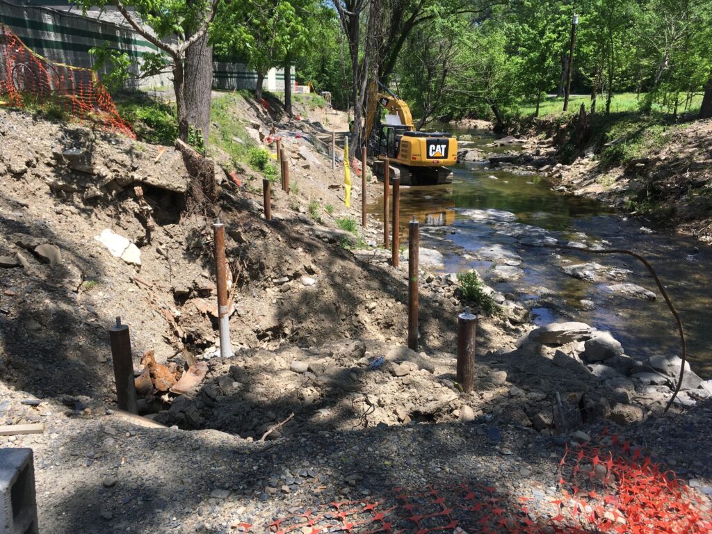 A construction site by a small stream, with an excavator and several metal posts in the ground. Orange safety netting is in the foreground, and trees with green foliage surround the area. Dirt and rocks are scattered around the site.