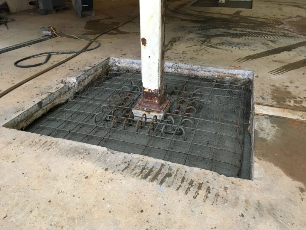 A concrete base with exposed rebar is set in a square cutout in the floor. A metal post is embedded in the center of the base. Construction tools and materials are visible in the background.