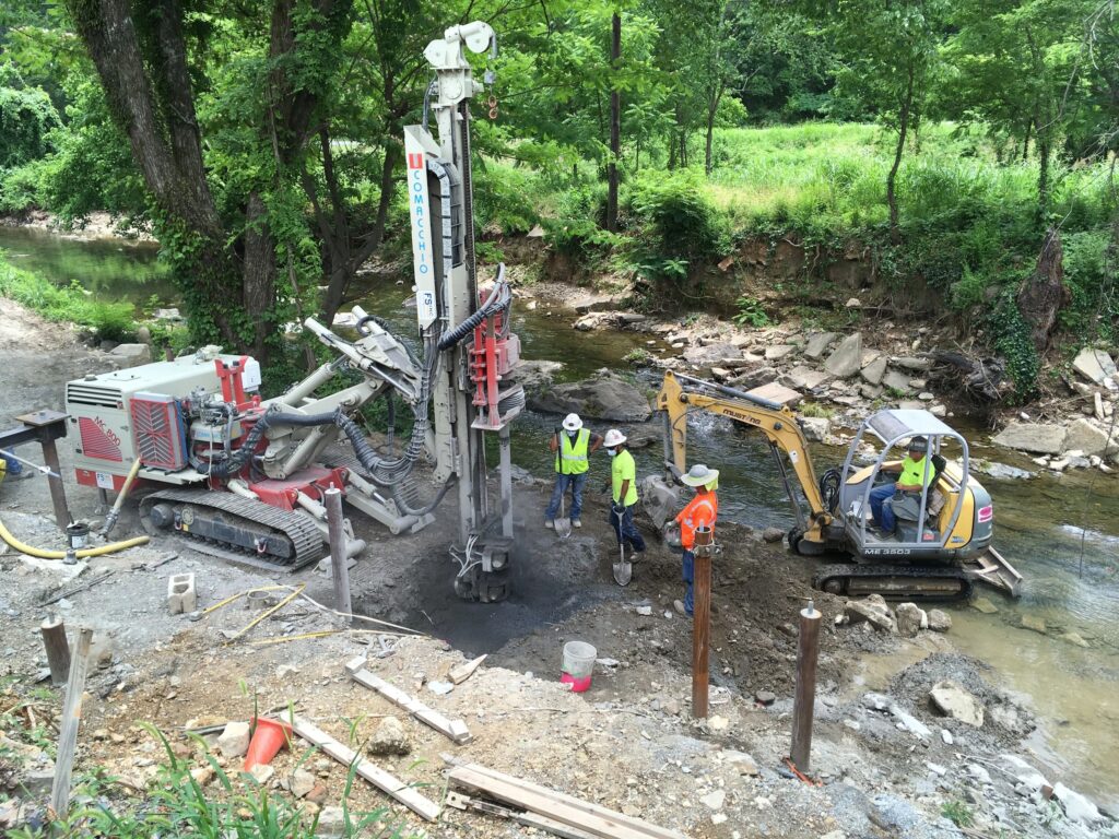 Construction workers operate machinery near a creek, surrounded by green trees. They use a drilling rig and mini excavator for soil sampling or foundation work. The scene is set in a natural, wooded area.