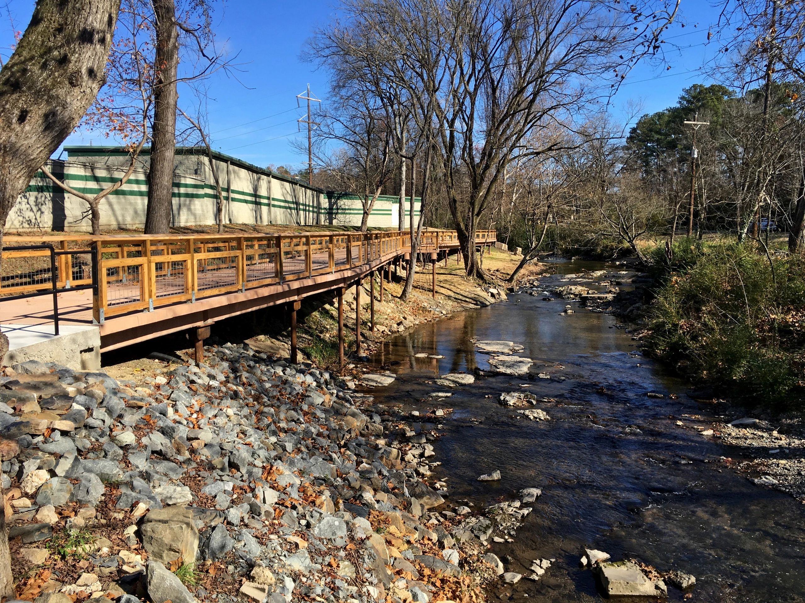 A winding creek flows alongside a wooden boardwalk elevated above a rocky bank. Bare trees line the path under a clear blue sky, and a building stands nearby. Fallen leaves scatter the landscape.