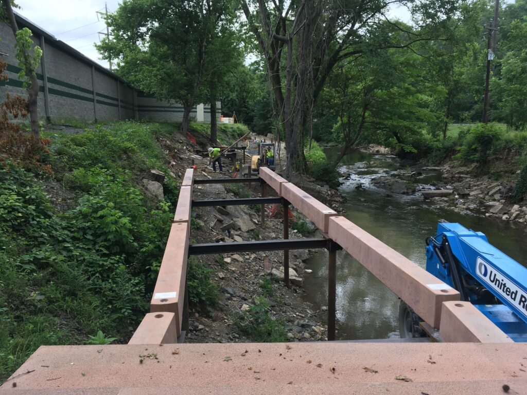 A construction site by a creek with workers installing beams. A blue lift vehicle is visible on the right, and a tall fence lines the left side. There are trees and greenery surrounding the site.