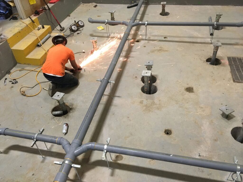 A worker in an orange shirt and helmet uses a tool to cut a pipe, sparking brightly. The pipes are arranged on a concrete floor with holes and supports, suggesting ongoing construction or installation work.