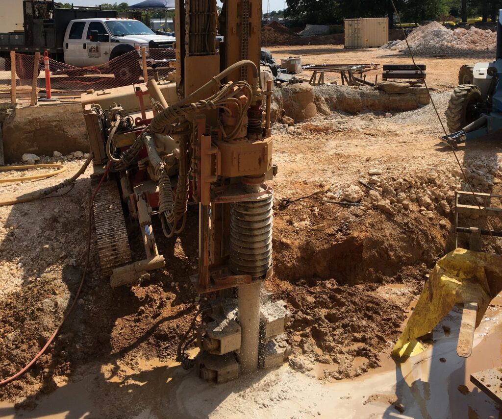 A large drilling machine operates at a construction site with loose soil and rocks. Nearby, a pickup truck and other equipment are visible. The site is partially cordoned off with orange safety fencing.