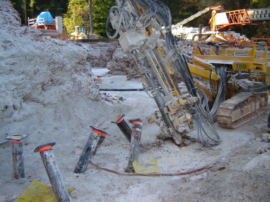 A construction site with a large yellow drilling machine positioned at an angle, surrounded by white debris. Several exposed pipes with red rims are visible amidst the dusty environment. Construction equipment is seen in the background.