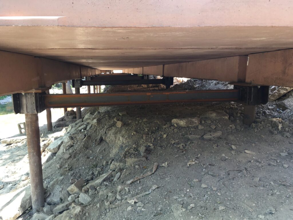 View of the underside of a bridge, showing concrete beams and metal supports. The ground below is covered with dirt, rocks, and debris. Sunlight filters through gaps, casting shadows underneath the structure.