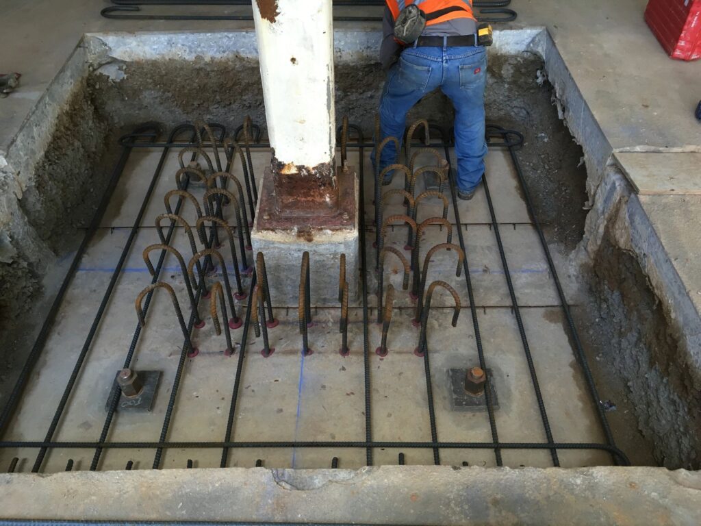 A construction worker wearing a high-visibility vest installs rebar around a concrete column base in a foundation pit. The rebar grid is neatly arranged, and anchor bolts are visible in the corners of the pit.