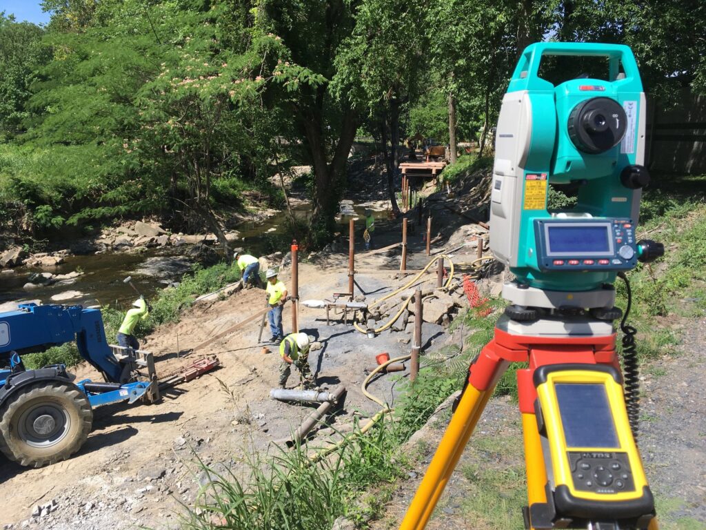 A construction site by a river with workers in safety gear setting up infrastructure. A green and white surveyor's instrument with a digital screen is in the foreground. Green trees and a partially completed structure are visible in the background.