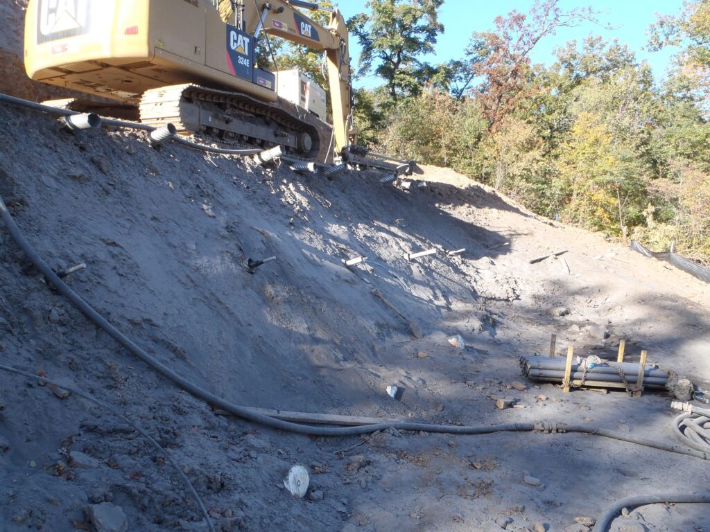 A construction site on a slope with a large excavator. The ground is uneven and dusty, with several pipes and hoses scattered around. There's a pile of materials in the foreground, and trees are visible in the background under a clear blue sky.