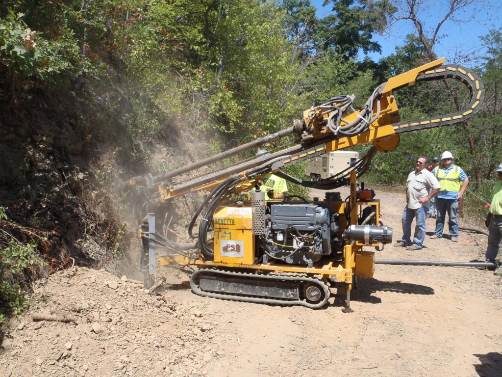 A yellow rock drilling machine operates on a dirt path surrounded by greenery. Dust is visible from the drilling. Several workers in hard hats and safety vests observe the process nearby.