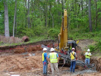 Four construction workers wearing hard hats and safety vests stand by a yellow excavator at a forested construction site. They appear to be discussing or overseeing excavation work amidst trees and earth-moving activity.