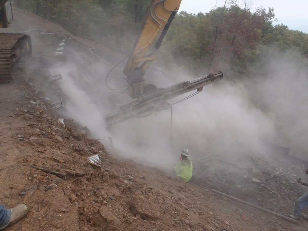 A construction site with workers operating heavy machinery on a dirt slope. Dust clouds are visible as a machine drills into the ground. Trees are in the background under an overcast sky.