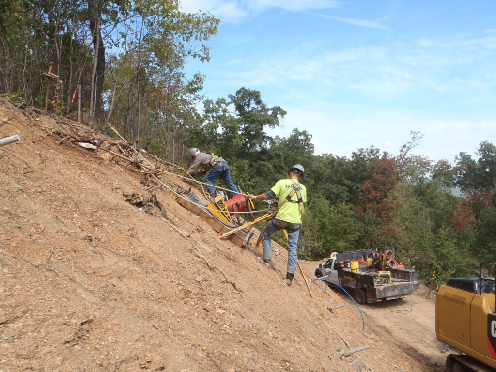 Two workers in safety gear perform maintenance on a steep, sandy hillside using ropes for support. They are surrounded by trees, with equipment and a truck nearby under a partly cloudy sky.