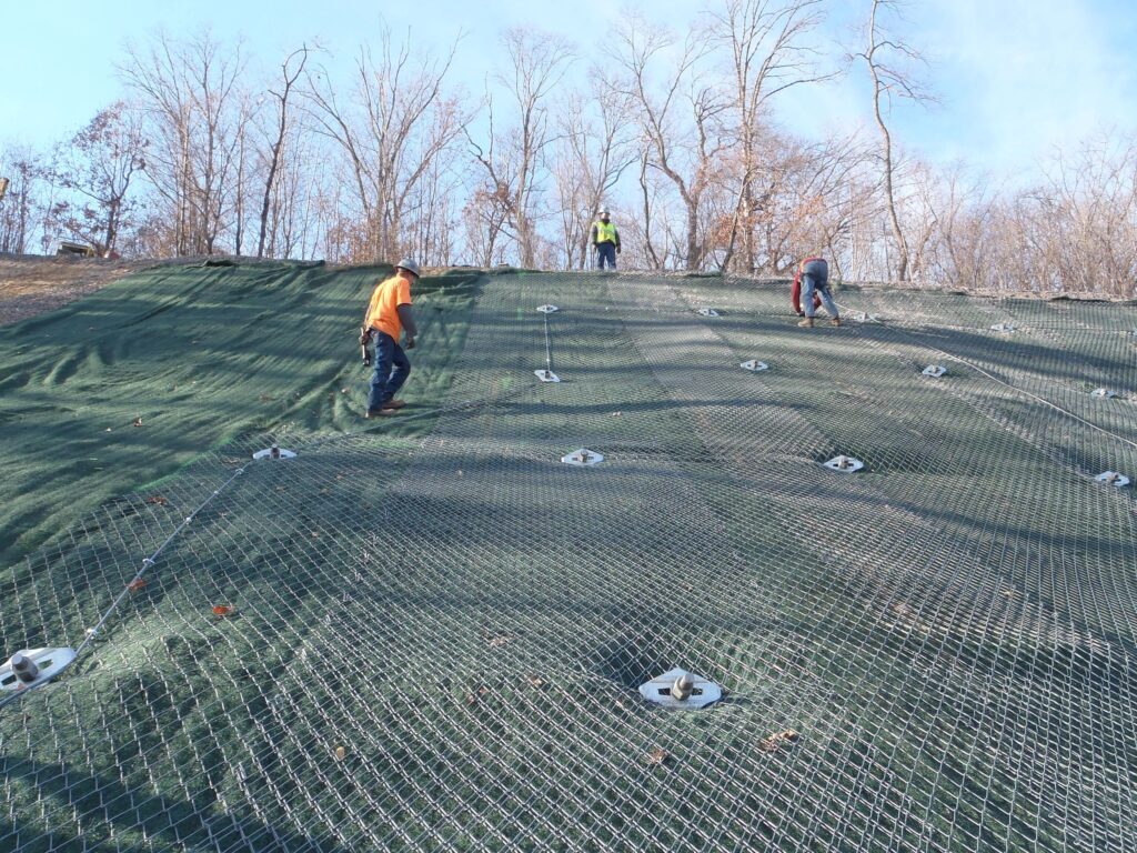 Workers secure a green net and wire mesh on a steep slope to prevent erosion. Bare trees stand in the background under a clear blue sky.