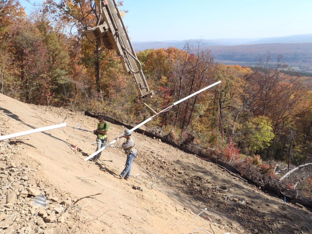 Two construction workers on a slope install a long white pipe with the help of machinery. The background showcases a forest with trees in autumn colors and distant hills under a clear sky.