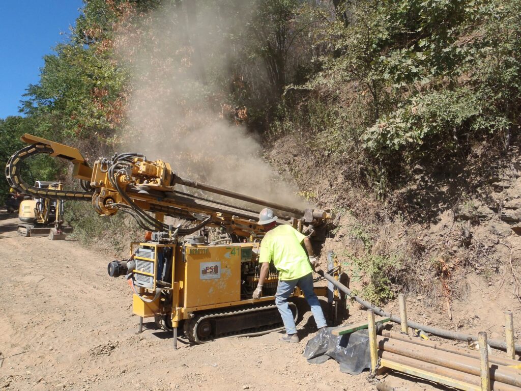 A worker operates a yellow drilling machine on a dirt road surrounded by trees. Dust rises from the machine as it drills into the hillside. A small excavator is visible in the background.