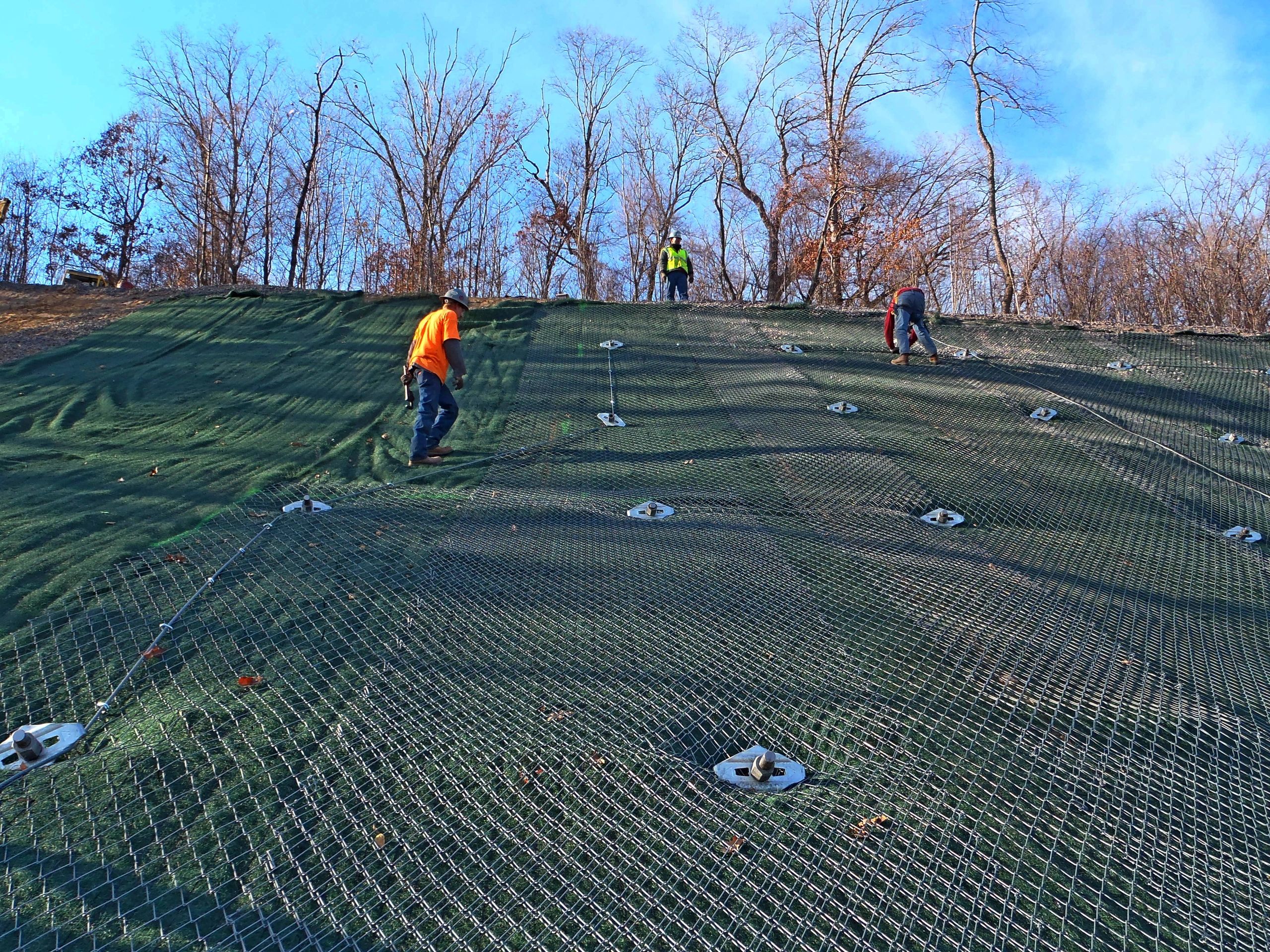 Three workers in orange and yellow vests secure a large wire mesh on a steep, grassy hillside surrounded by bare trees. The sky is clear and blue, and the landscape indicates a construction or erosion control project.