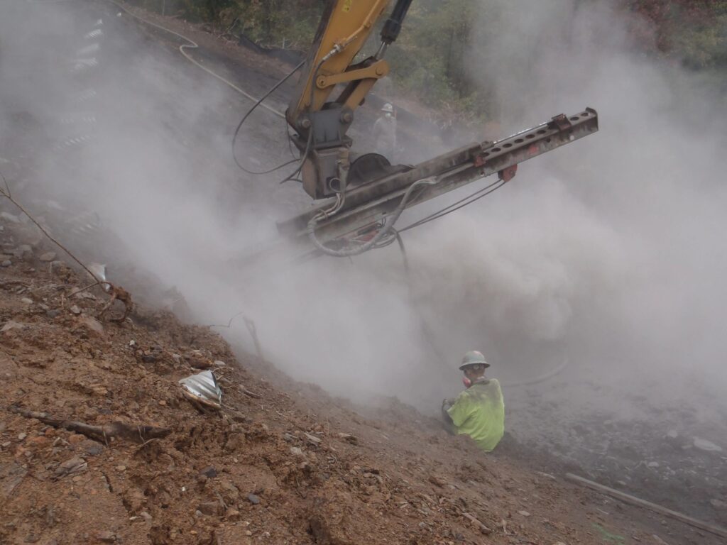 A construction worker in a yellow safety jacket and helmet sits beside a large, smoke-emitting machine on a rocky site. The area is filled with fog-like smoke, and there's a slope of dirt in the foreground.