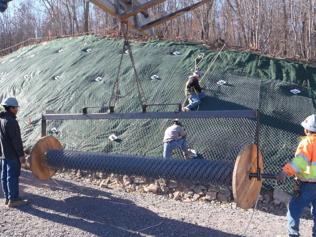 Workers install netting on a sloped, vegetated hill using a large spool of wire mesh. The scene includes several crew members wearing safety gear and helmets, with trees in the background on a clear day.