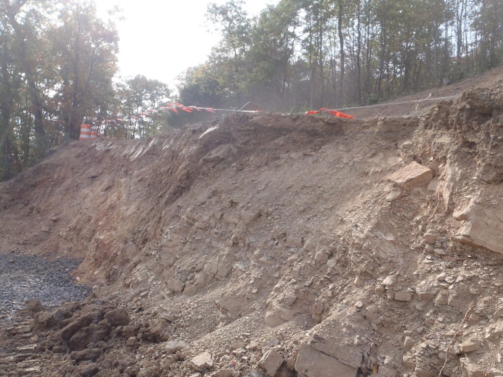 Steep rocky slope with exposed earth and large cracks. A row of bright orange safety cones and tape is placed along the top edge. Trees can be seen in the background under a cloudy sky.