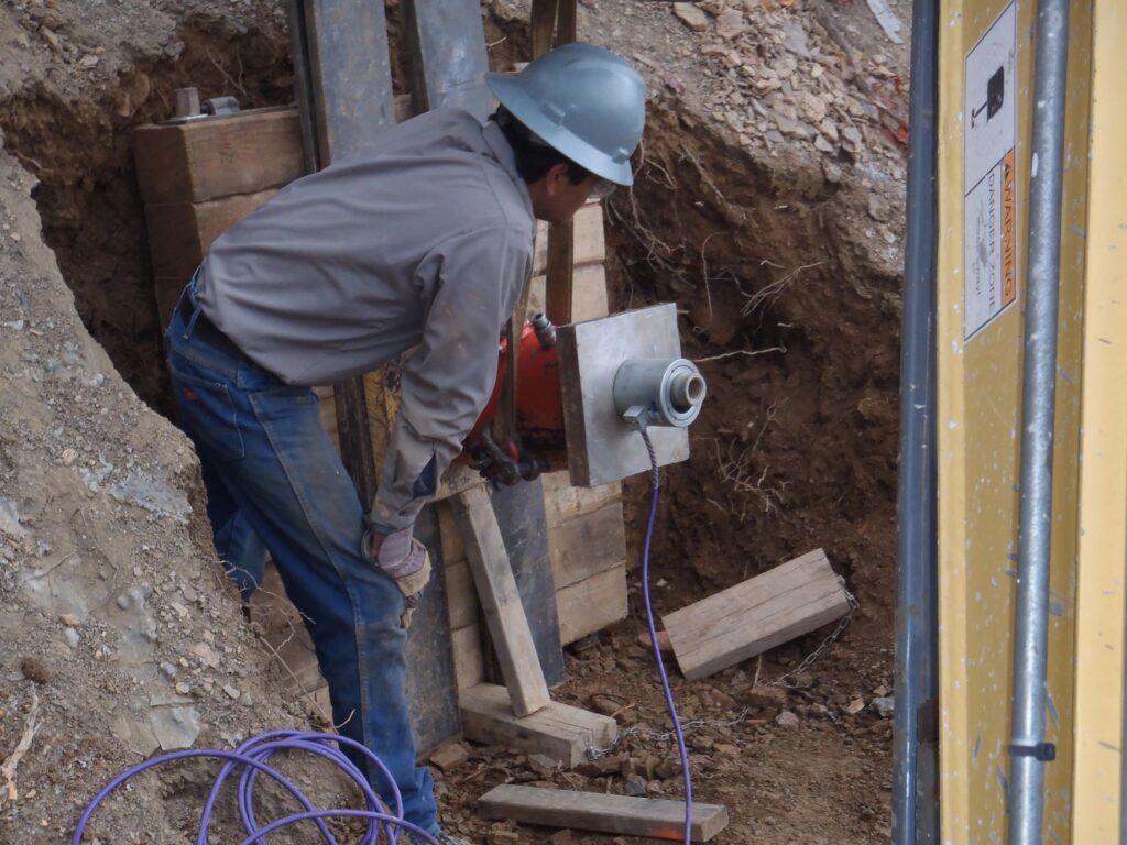 A construction worker wearing a hard hat and gloves operates machinery at a construction site. The worker is bending forward, focusing on the equipment, with wooden beams and earth around him. A yellow structure partially frames the right side.