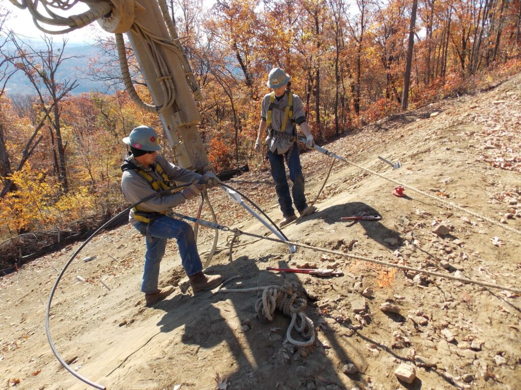 Two workers in safety gear and helmets are on a wooded hillside, using cables and ropes for equipment maneuvering. The setting is an autumn forest with colorful leaves. They appear focused and are wearing harnesses for safety.