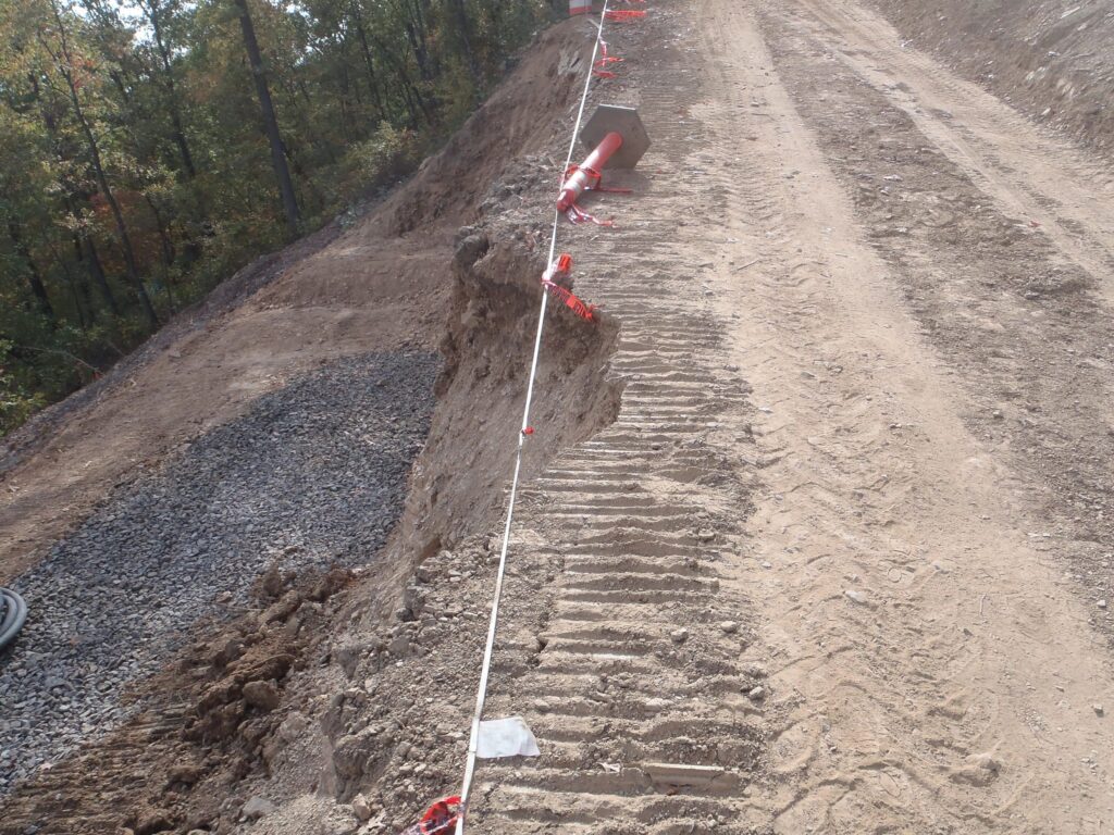 A dirt road under construction with visible tire tracks runs alongside a steep drop-off, marked by red and white striped tape for safety. The area is bordered by trees on the left, and gravel fills part of the slope.