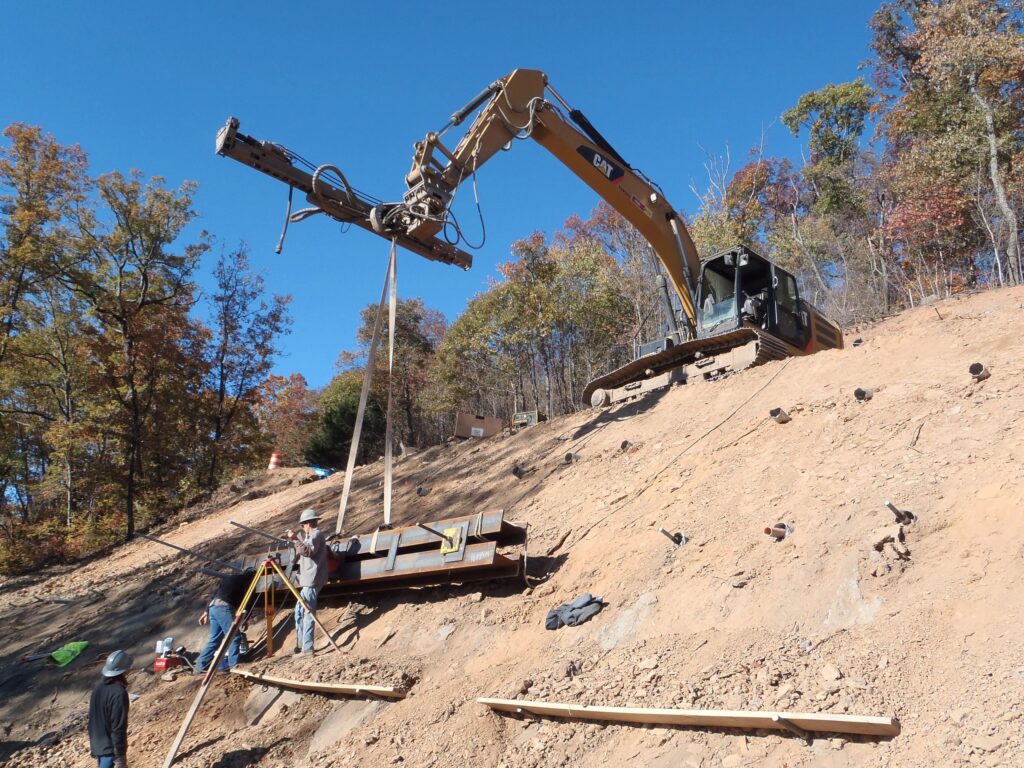 Construction workers are using an excavator with a long attachment on a sloped, dirt-covered hillside. The area is surrounded by trees with autumn foliage under a clear blue sky.