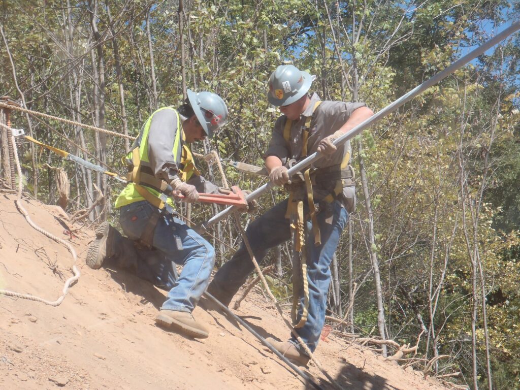 Two construction workers wearing helmets and safety gear install a metal pole on a sloped, sandy terrain. They are surrounded by trees and sunlight, focusing intently on their task.