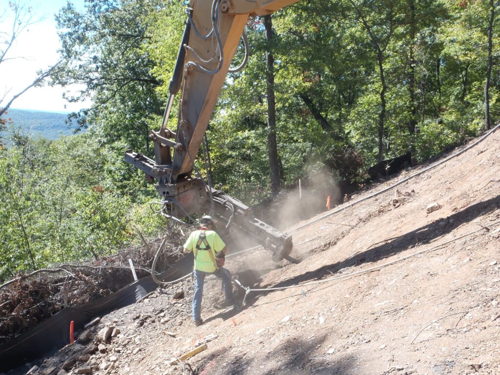 A construction worker operates heavy machinery on a steep, wooded hillside. Dust is rising from the ground as the machine digs into the slope. Trees surround the area, and a barrier runs along the hillside. The sky is clear, suggesting a sunny day.