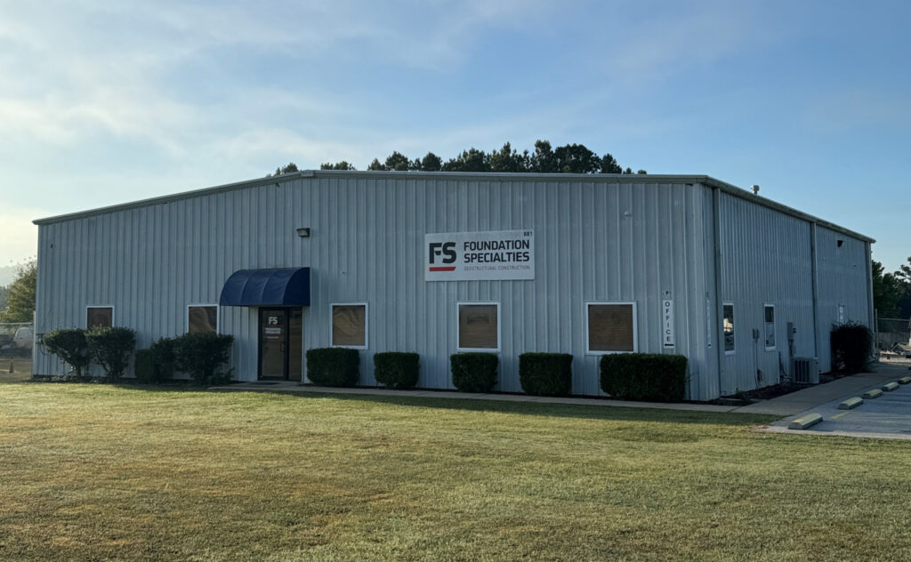 A large steel building with a sign "FS Foundation Specialties" on the front. The structure has a blue awning over the entrance and is surrounded by trimmed bushes. The building sits on a grassy lawn under a clear sky.