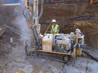 A construction worker wearing a helmet and safety vest operates a drilling machine at a construction site. The area is surrounded by dirt and various tools, with a partially constructed building in the background.